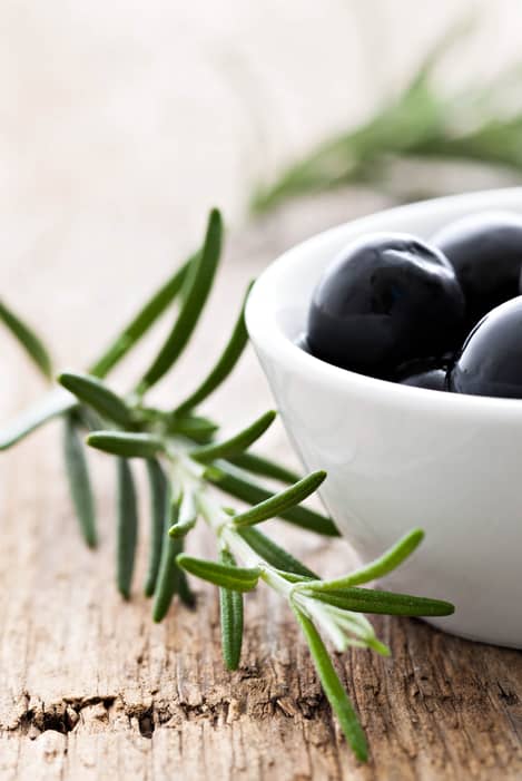 Close up of black olives in a small white bowl on a wooden background with a sprig of rosemary in the foreground.