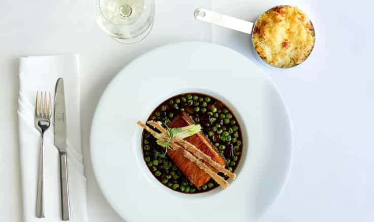 Over-head shot of table setting with white table cloth; wide-rimmed, white plate with stylish fish dish in it; wine glass, small saucepan with toasted mashed potato as sides.