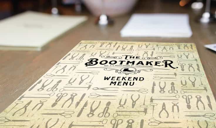 The Bootmaker restaurant menu design cover placed on a dark wooden table with place setting out of focus in background.