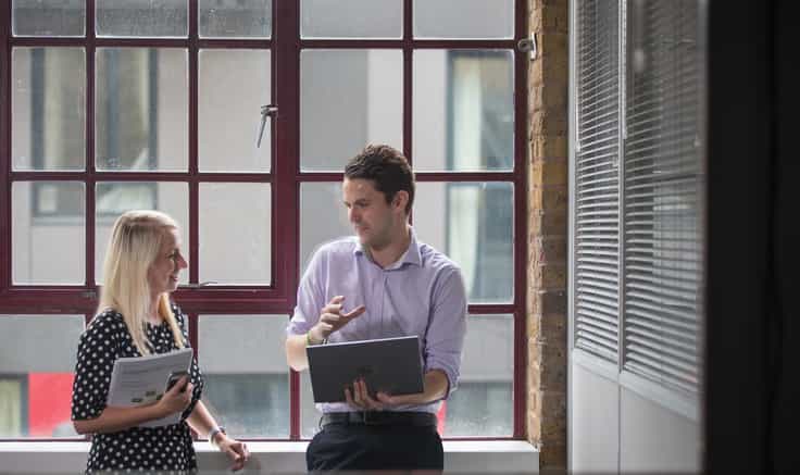 A man and woman dressed smartly, stood in front of a window of an industrial style office room.