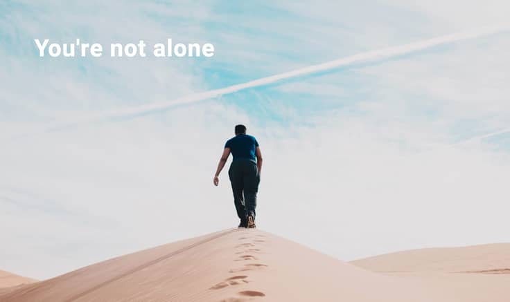 Man in distance walking through desert dunes with trail of footsteps in the sand. Blue sky in background.