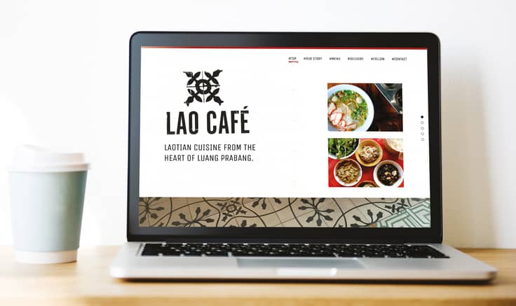 Laptop on plain wooden table top with pale blue take-away coffee cup to its side. The laptop is displaying the Lao Cafe homepage on its screen which features a retro-style logo and square images Laotian food in simple bowls.