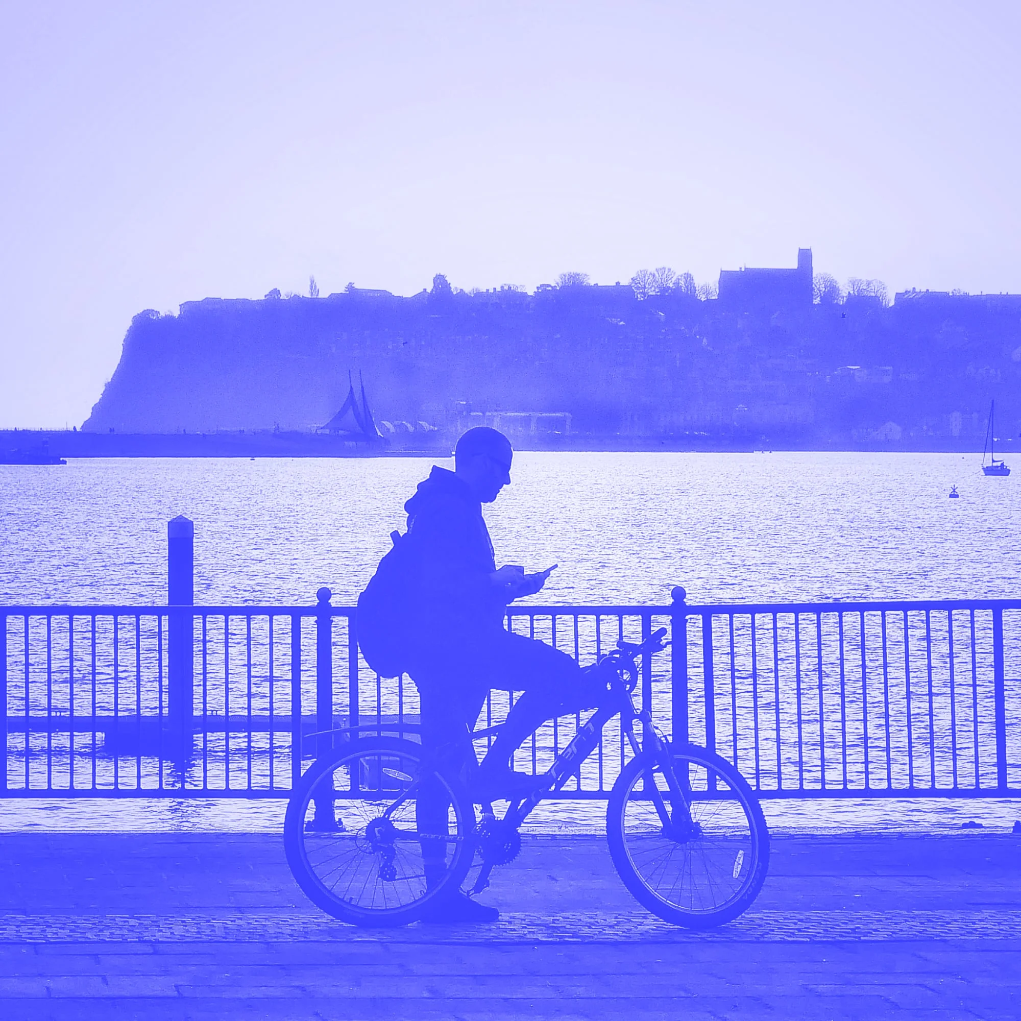 silhouette of man on bicycle checking phone by waterfront blue toned