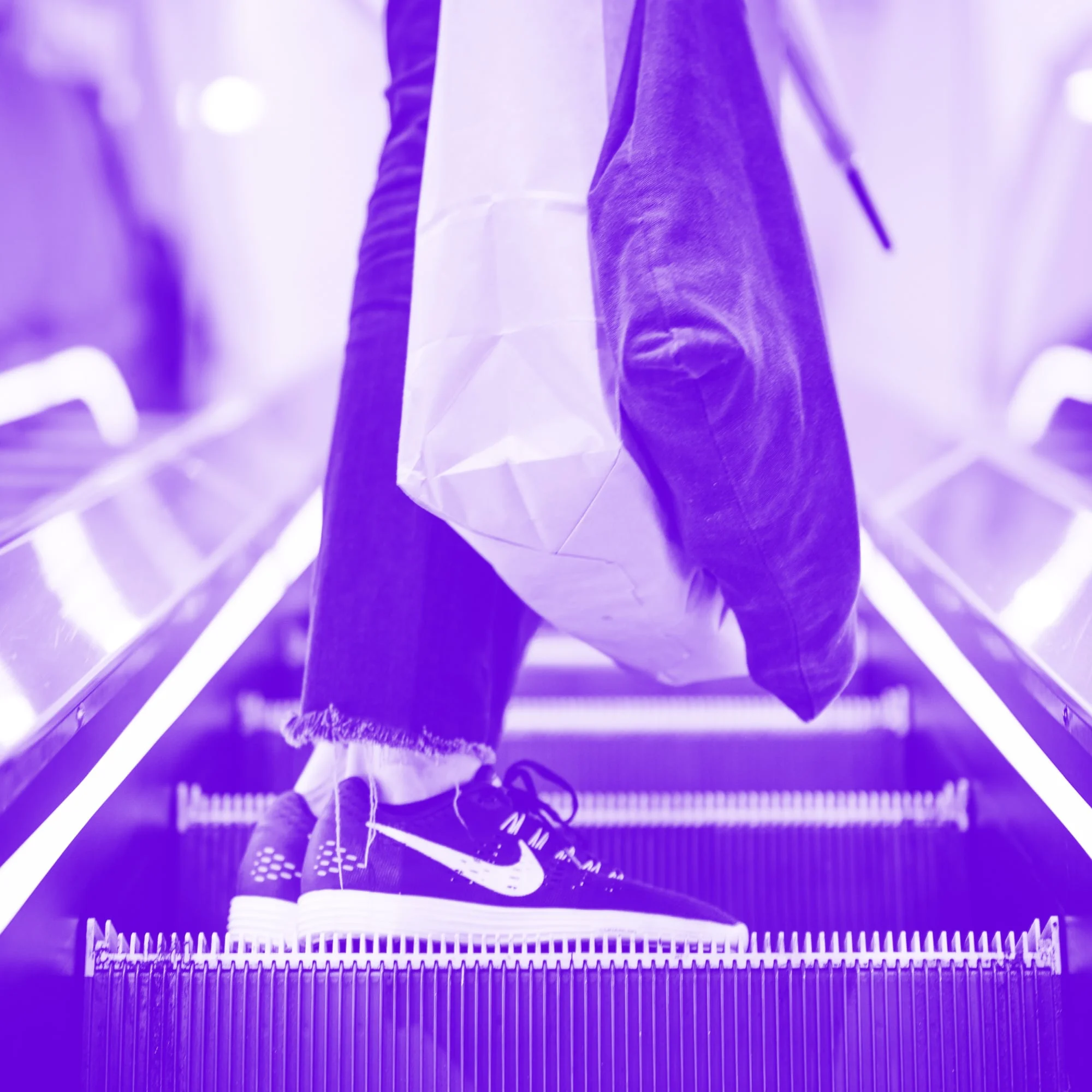persons feet in nike trainers on escalator holding white shopping bag purple toned