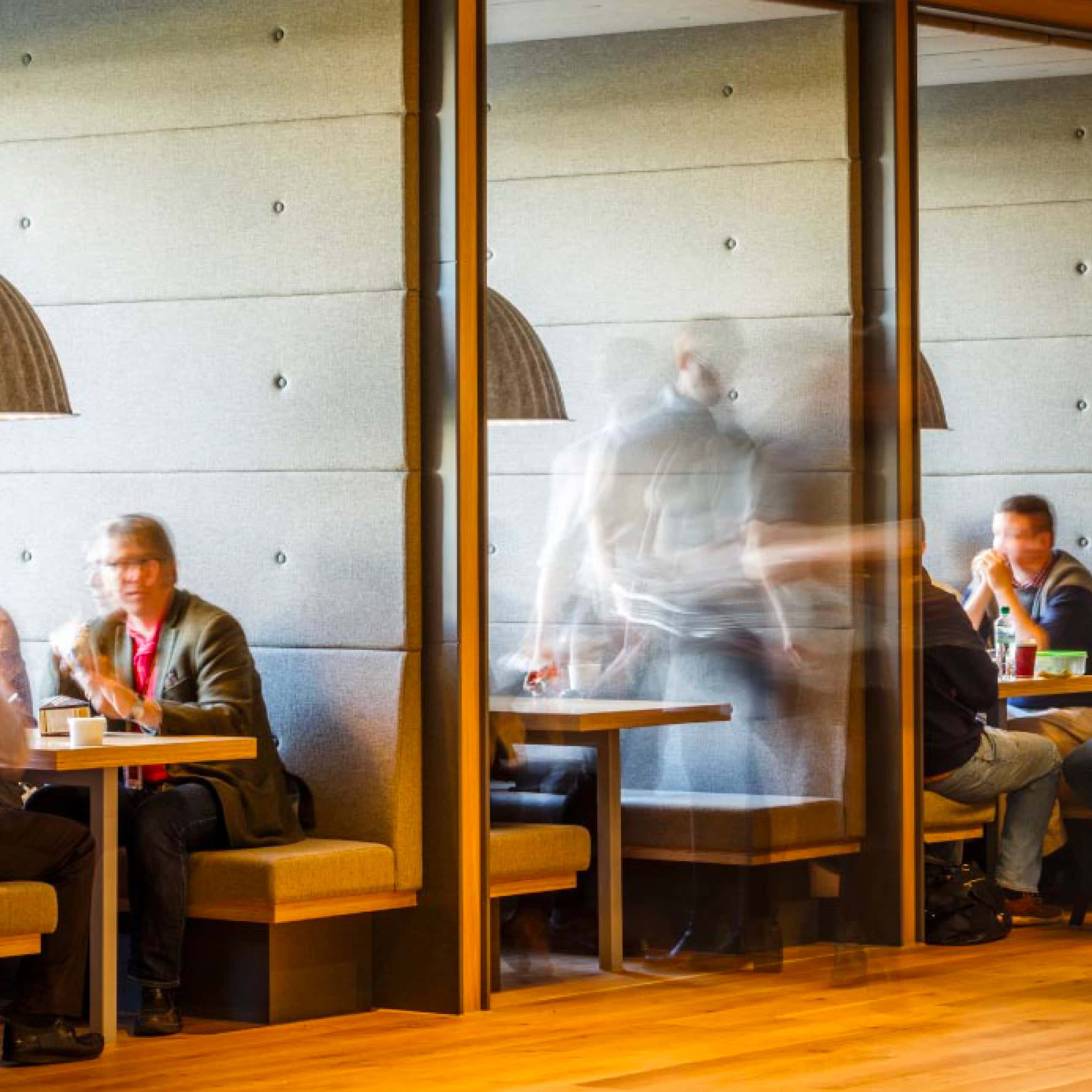 People seated in office café booths with motion blur behind glass partitions