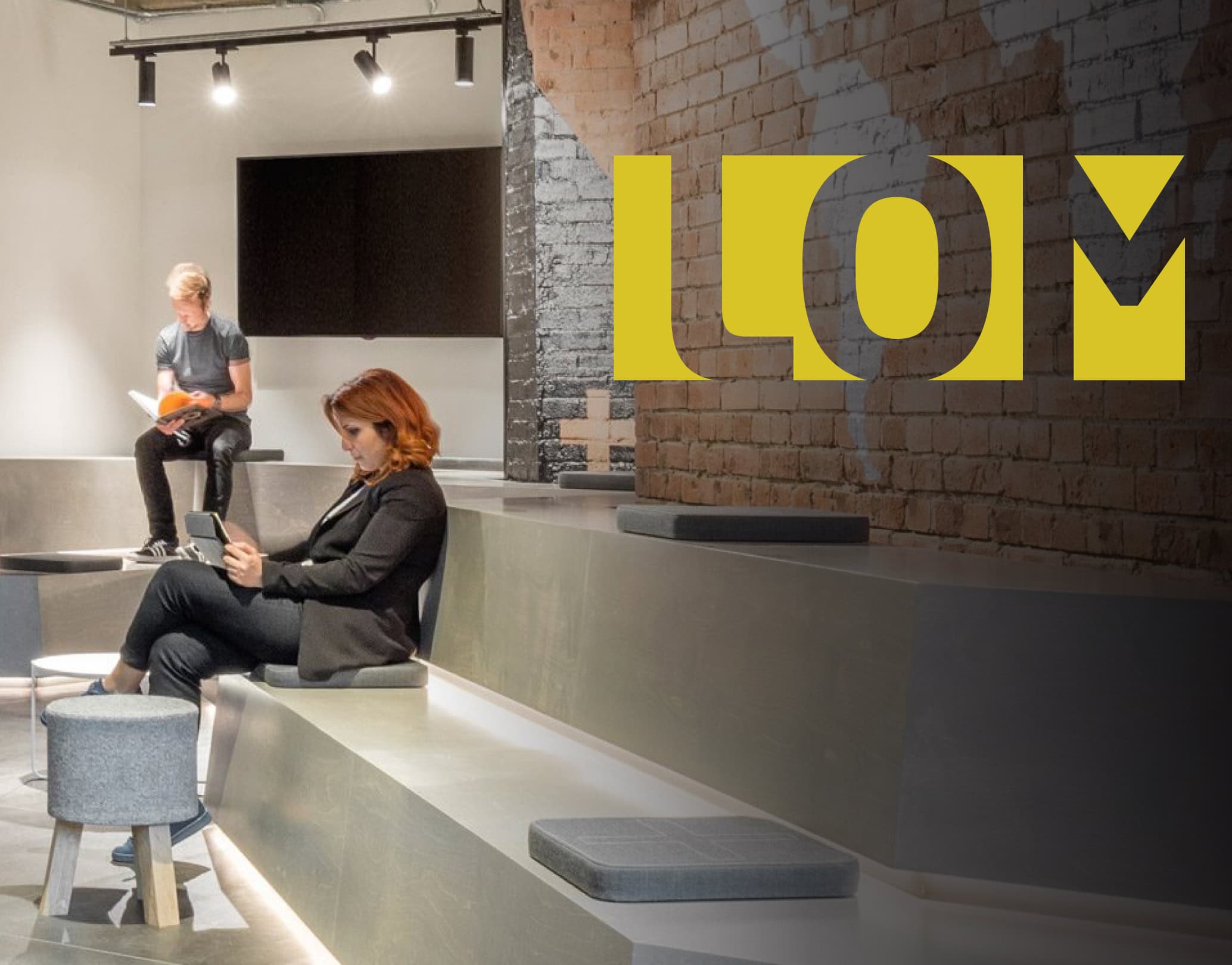 Modern studio lounge with tiered concrete seating, two people working, and a large yellow LOM logo on a brick wall.