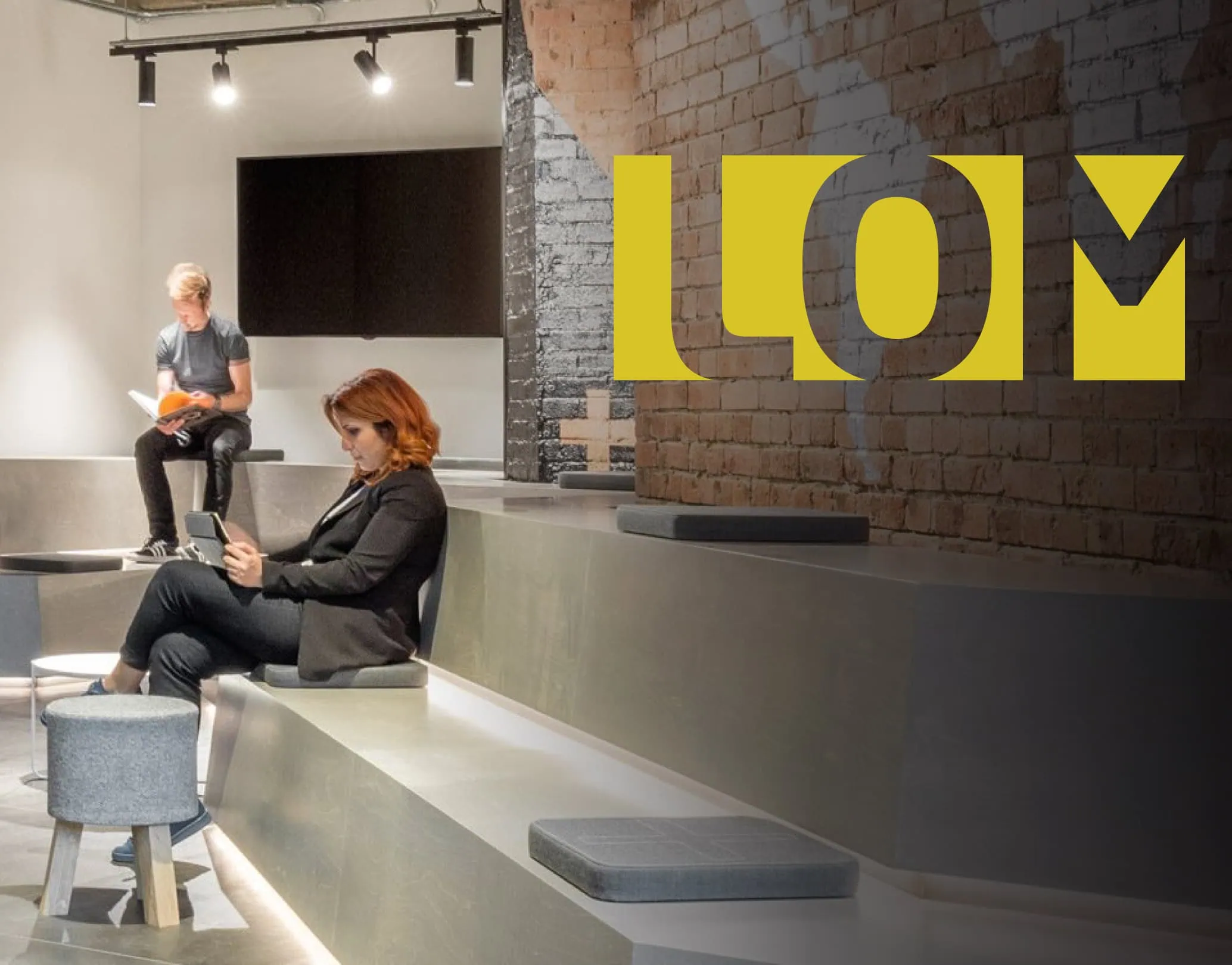 Modern studio lounge with tiered concrete seating, two people working, and a large yellow LOM logo on a brick wall.
