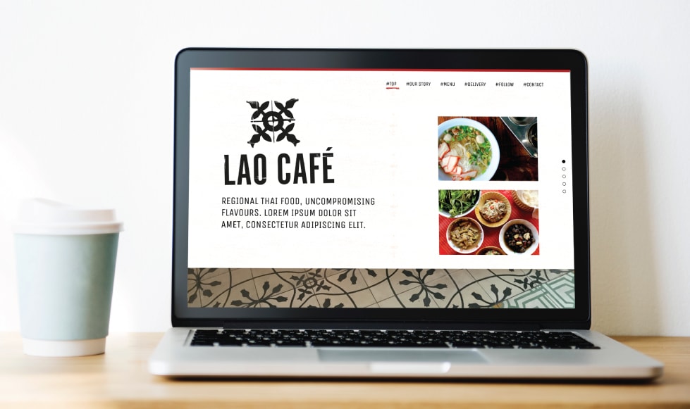 Lap top computer on wooden table with plain white backgroundna dn a pale blue take-out coffee cup. Lao Cafe one-page website template displayed on the screen.