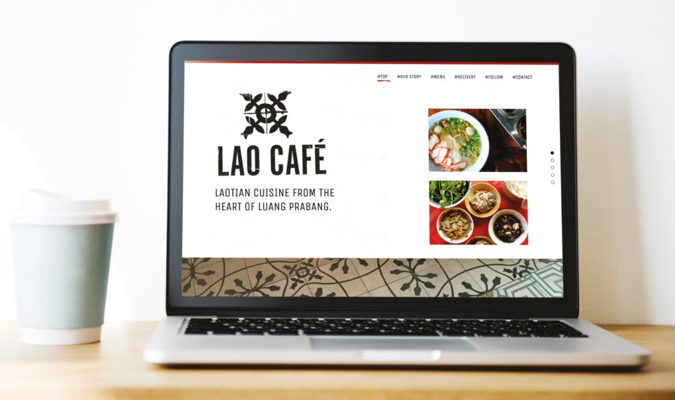 Laptop on plain wooden table top with pale blue take-away coffee cup to its side. The laptop is displaying the Lao Cafe homepage on its screen which features a retro-style logo and square images Laotian food in simple bowls.