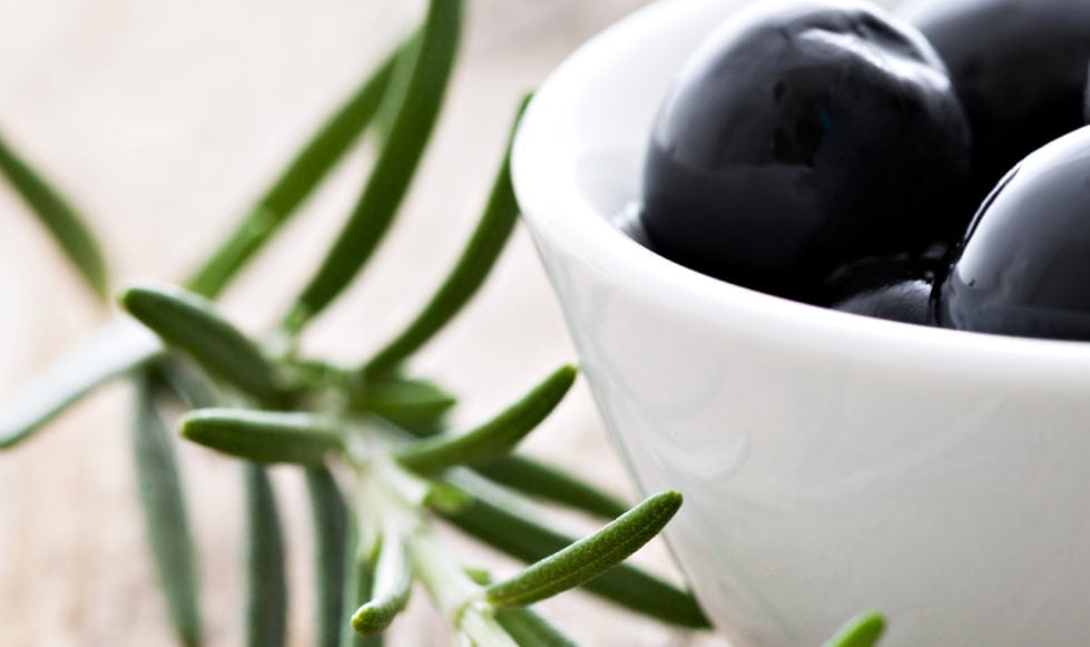 Close up of black olives in a small white bowl on a wooden background with a sprig of rosemary in the foreground.