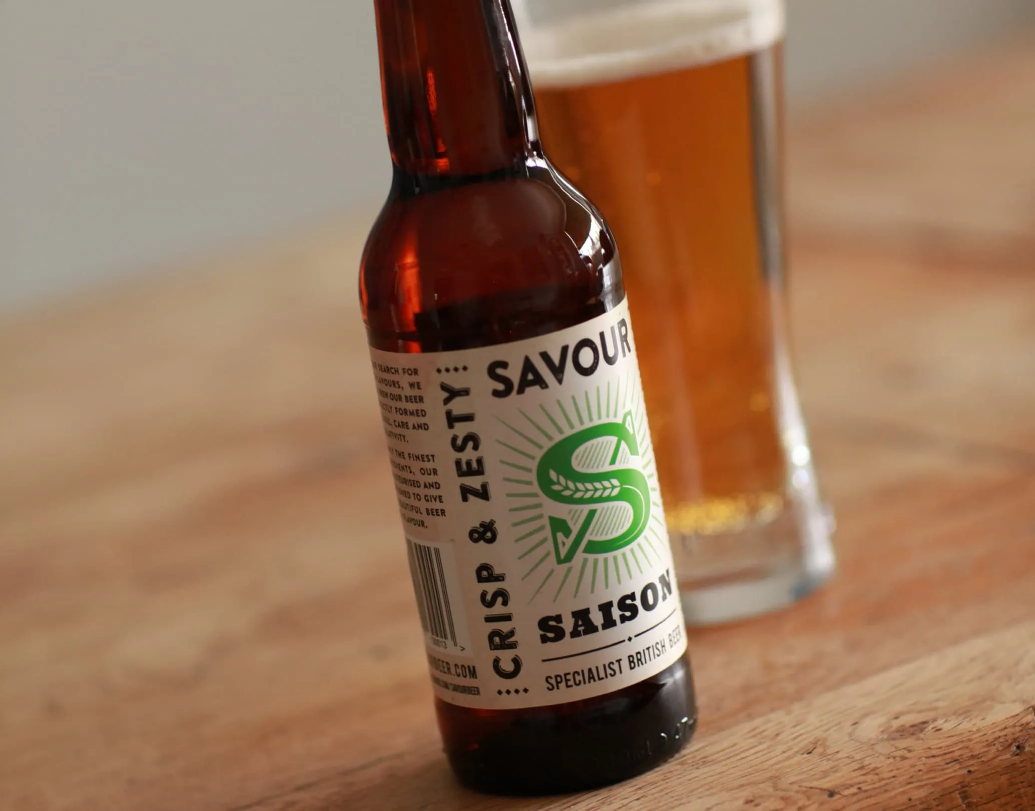 Savour Saison beer bottle on a wooden table with a glass of beer in the background