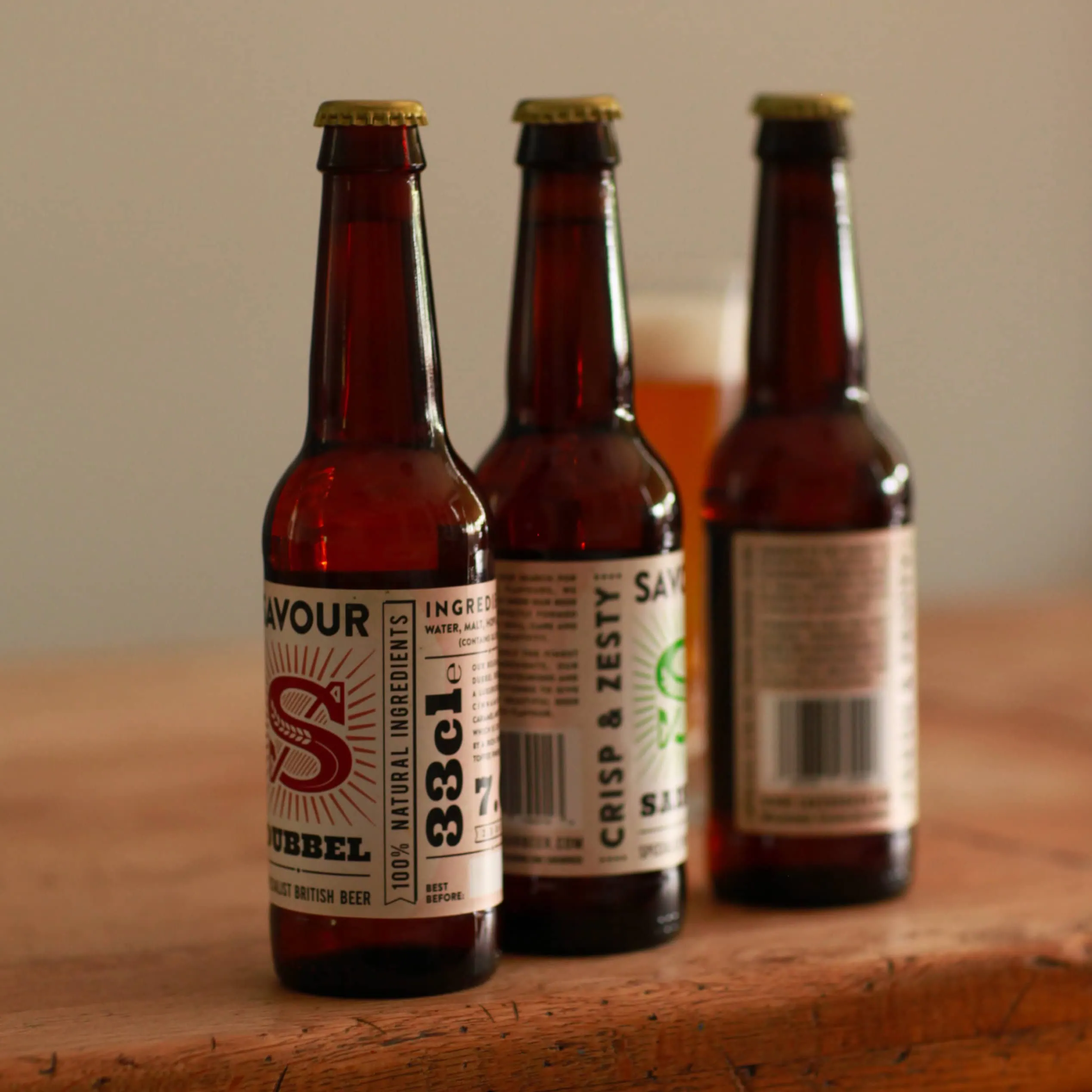Three Savour beer bottles on a wooden table with a glass of beer in the background