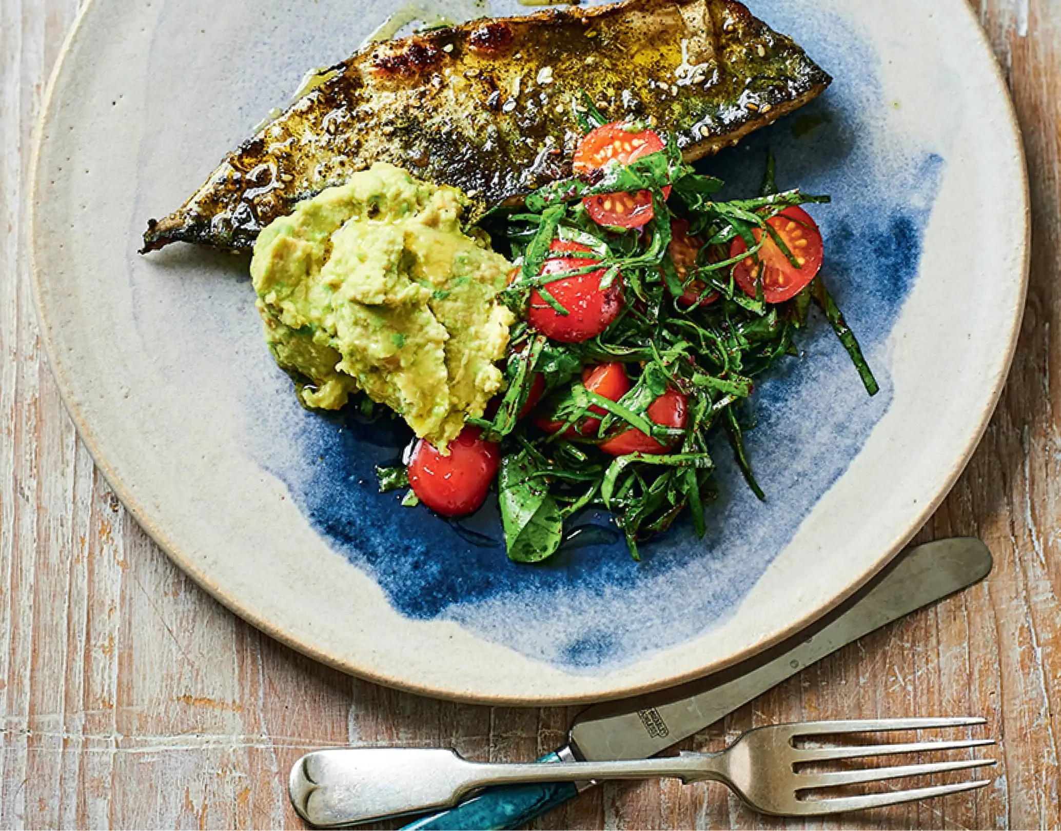 Grilled fish fillet with smashed avocado and cherry tomato salad on a blue-rimmed ceramic plate.