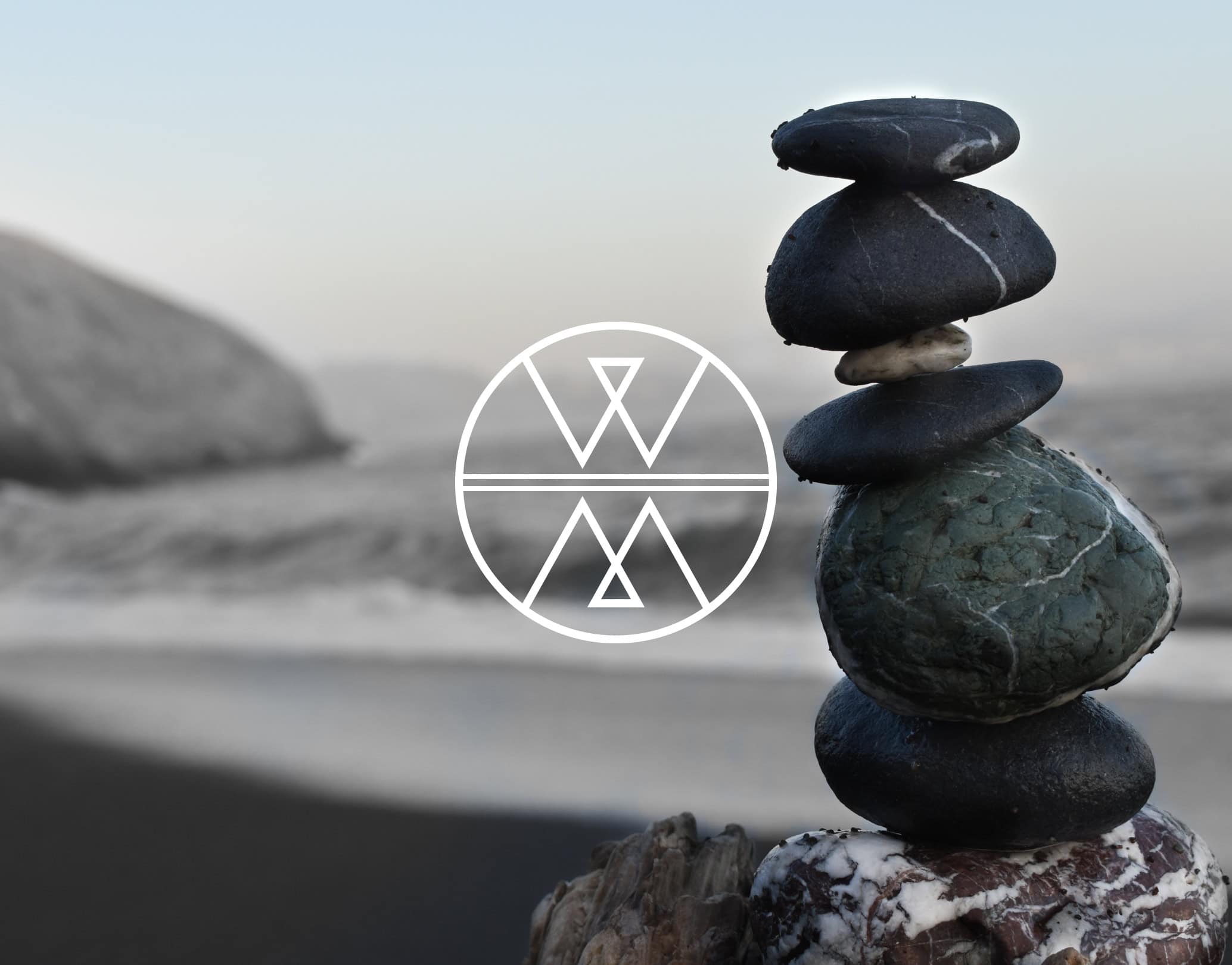 Stacked smooth beach stones on the shoreline with a white Wolf & Mermaid logo overlay
