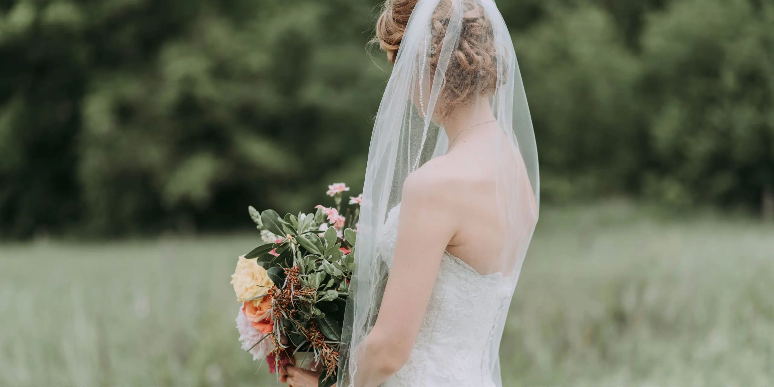Bride in a lace dress and veil holding a bouquet in a meadow