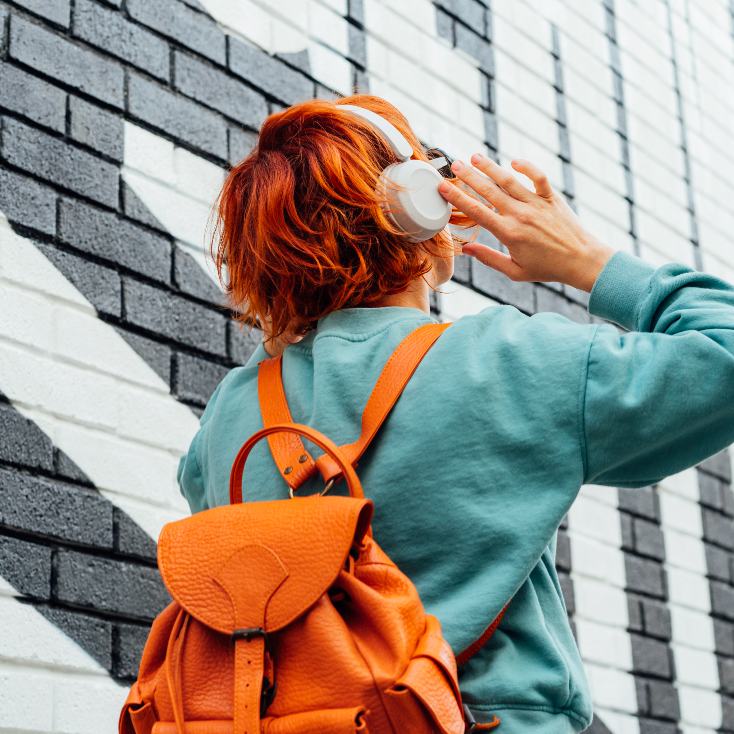 Red-haired person with orange backpack adjusting headphones beside a patterned brick wall