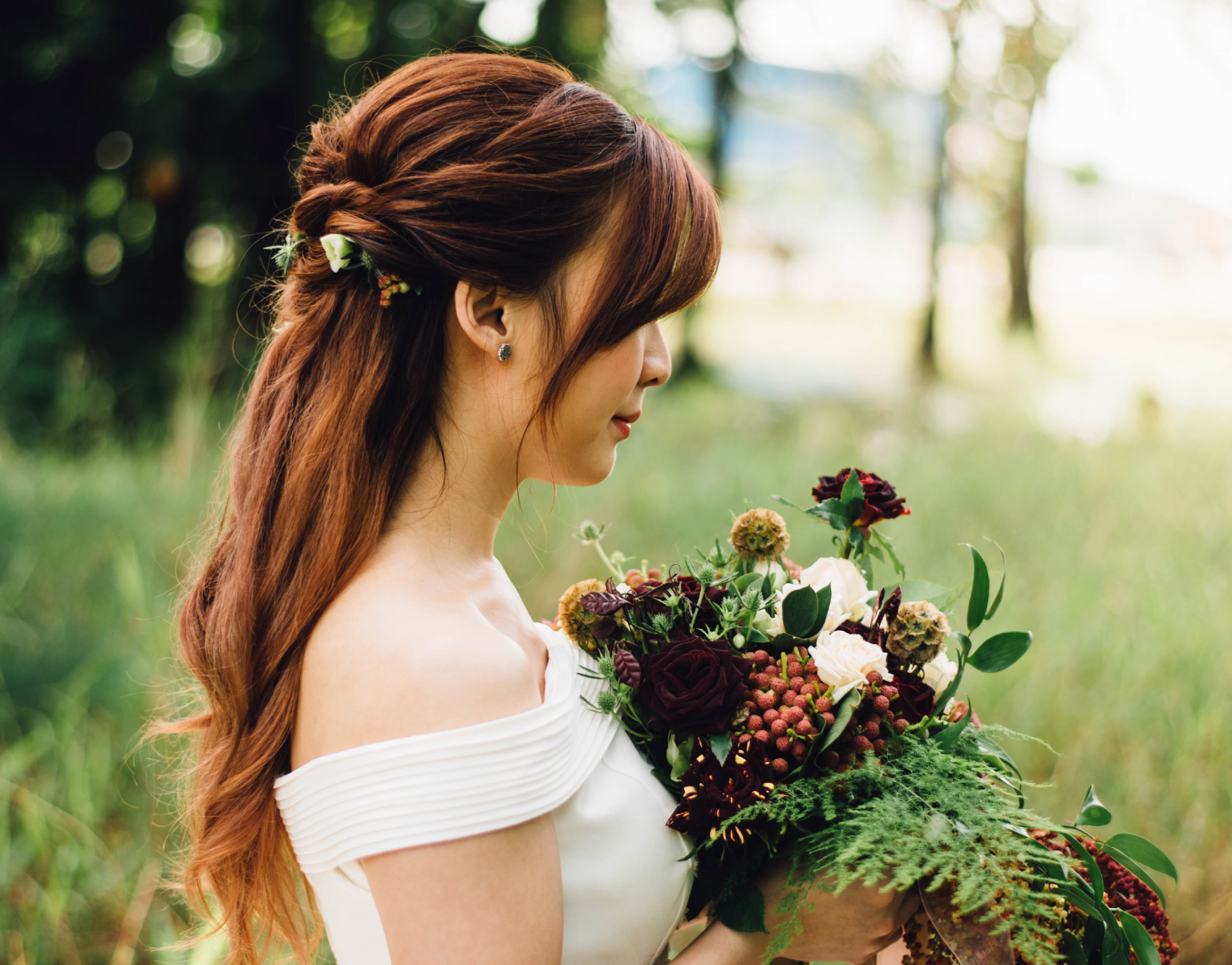 Bride in profile with long auburn hair holding a bouquet of dark red and white flowers outdoors in soft, dappled light.