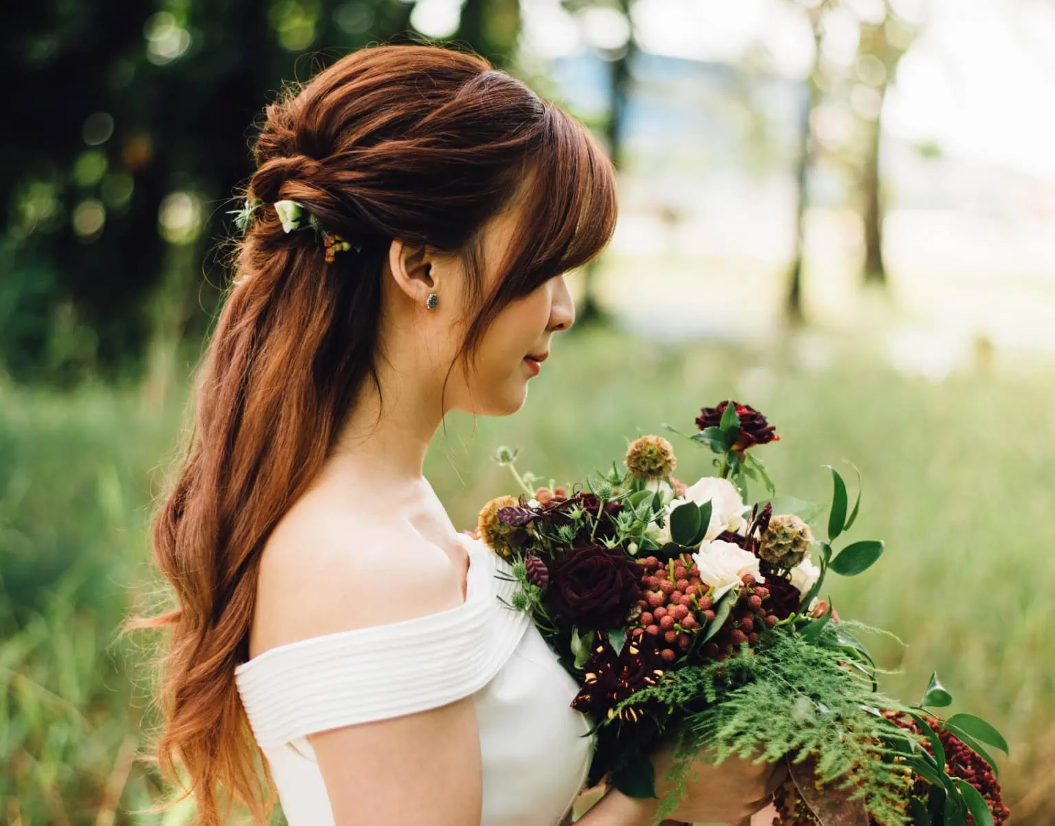 Bride in profile with long auburn hair holding a bouquet of dark red and white flowers outdoors in soft, dappled light.