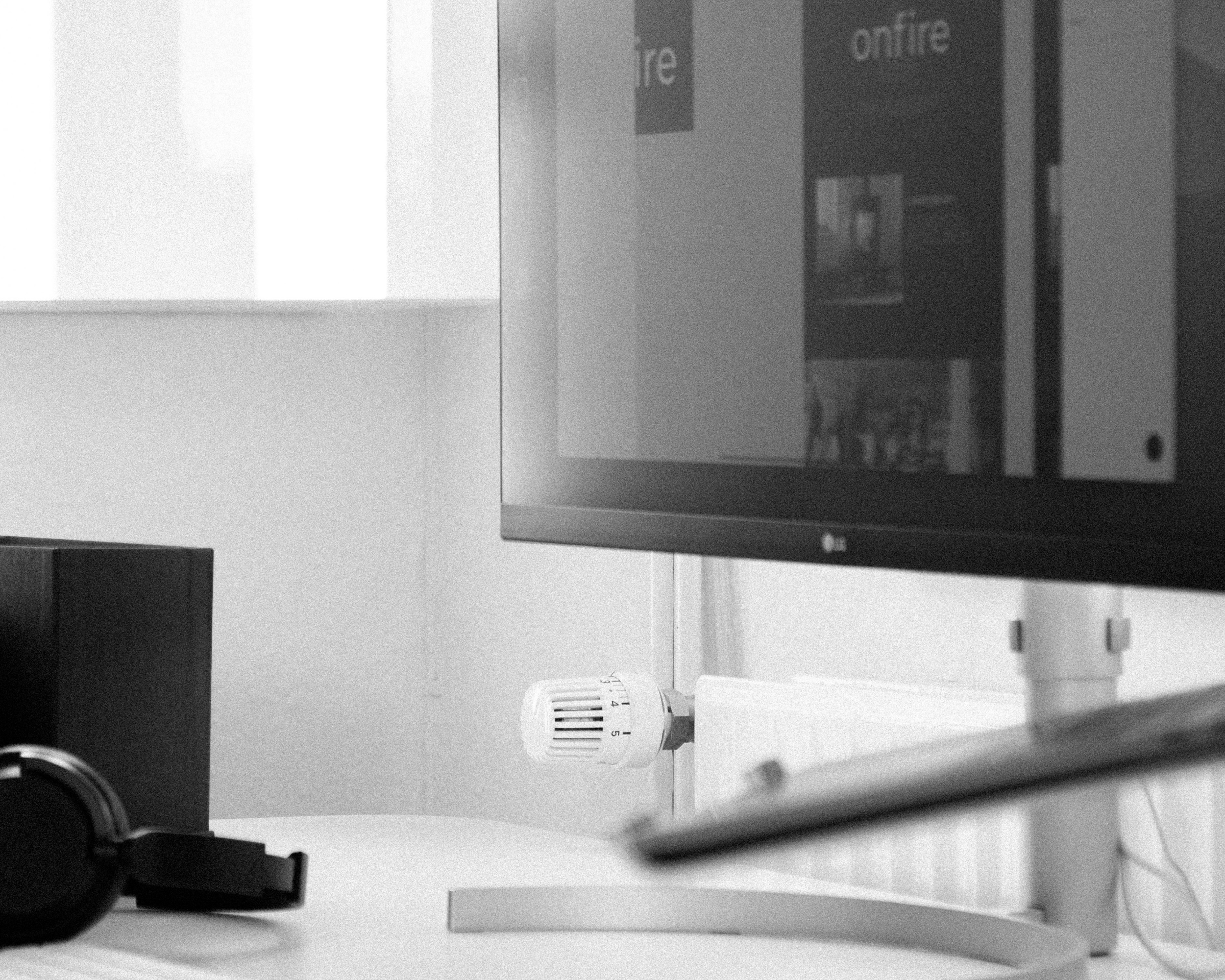 Black-and-white close-up of a desk with headphones, a computer monitor showing a website, and a radiator thermostat in the background.