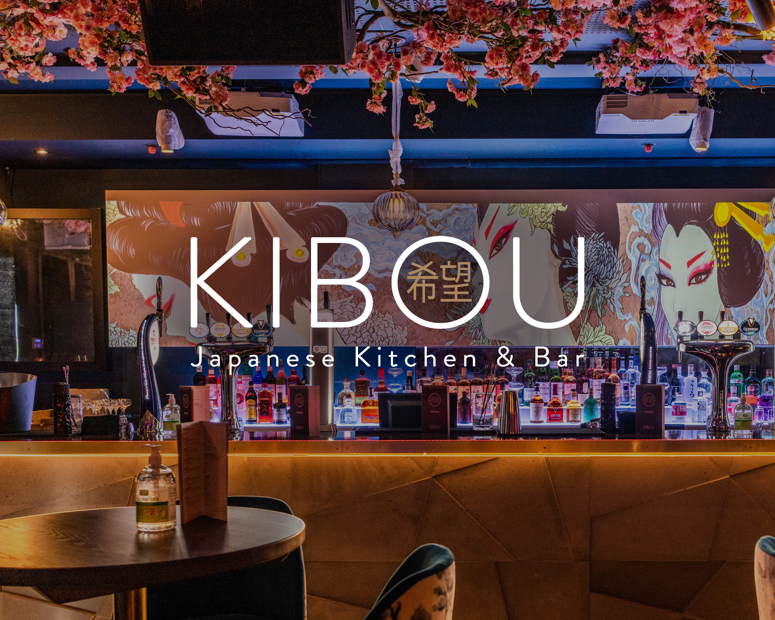 Interior of Kibou Japanese Kitchen & Bar, featuring a moody, atmospheric bar area with warm amber lighting. The back wall displays bold Japanese-style illustrated artwork including a kabuki-masked figure. Shelves are lined with spirits and bottles, pink cherry blossom branches hang from a dark ceiling, and the Kibou logo with Japanese characters is overlaid in white across the centre of the image.