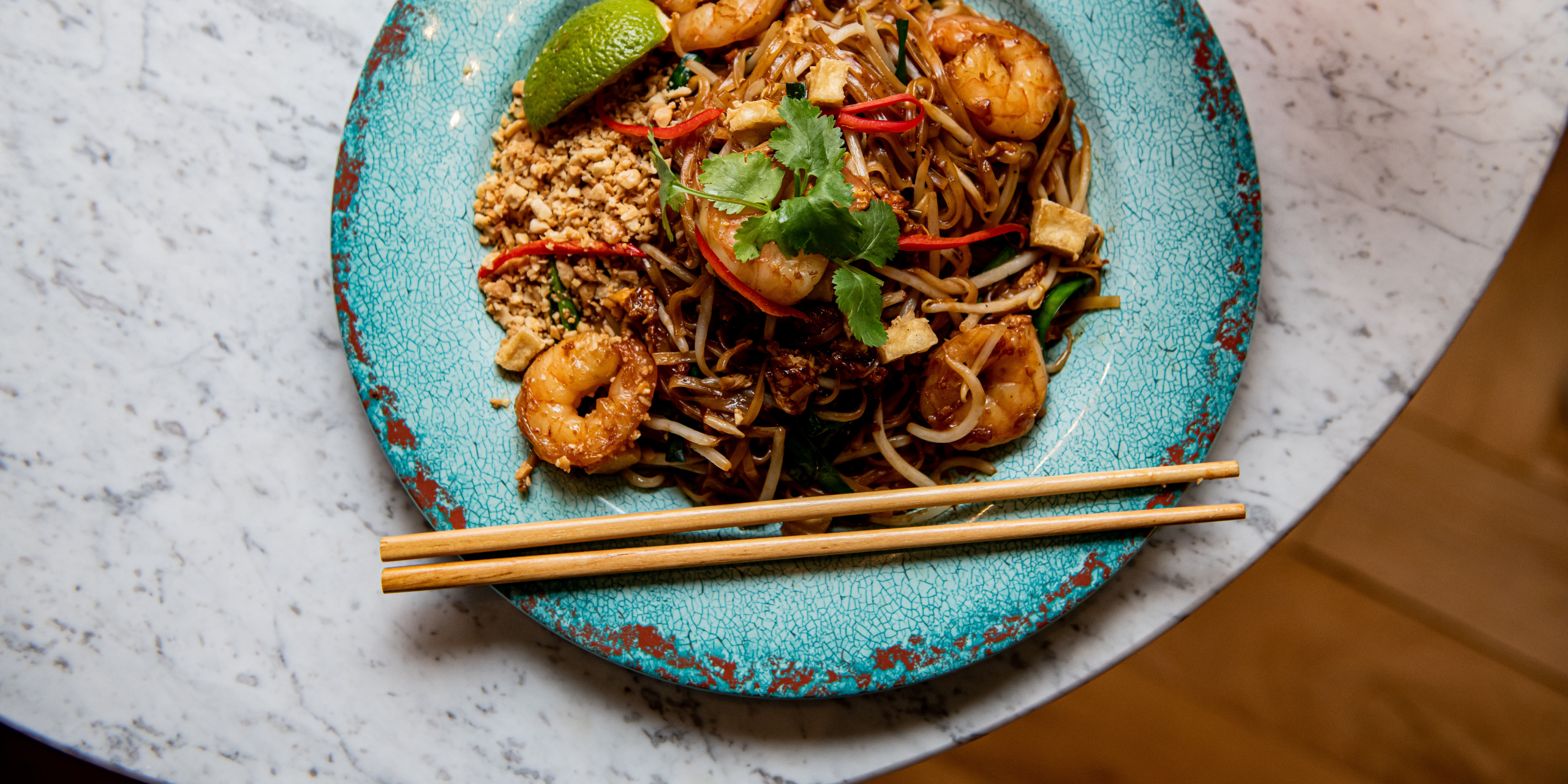 Overhead shot of a prawn Pad Thai served on a rustic turquoise ceramic plate, topped with fresh coriander, sliced red chilli, crushed peanuts, tofu, bean sprouts, and a wedge of lime. Wooden chopsticks rest across the front of the plate, set on a white marble table.